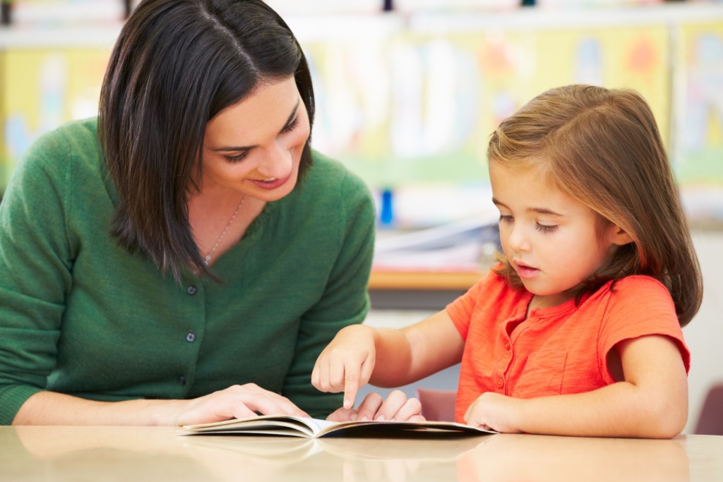 Teacher with pre-school girl at desk reading together