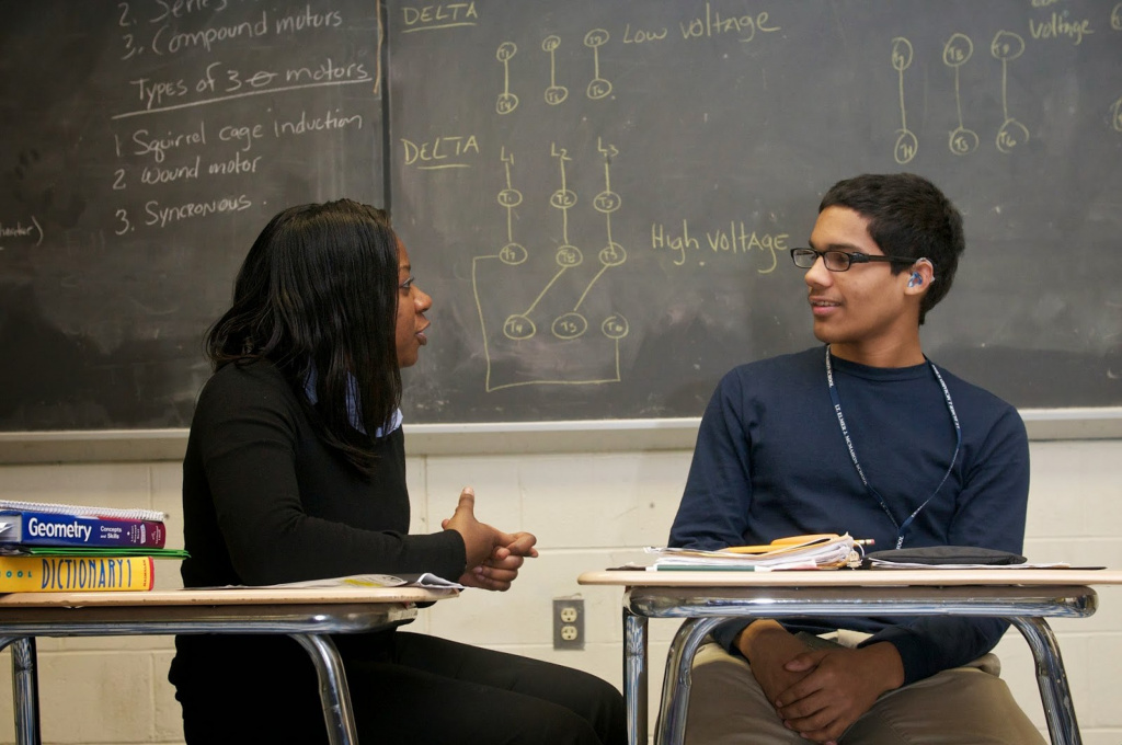 Two young adults, one with hearing aid, sitting at desks talking