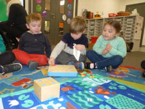 Children sitting round mat and looking at small wooden cube robot