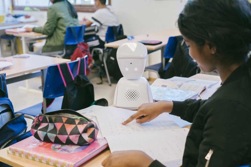 Girl sitting at a school desk with books and a pencil case. there is also an AV1 robot sitting on the table