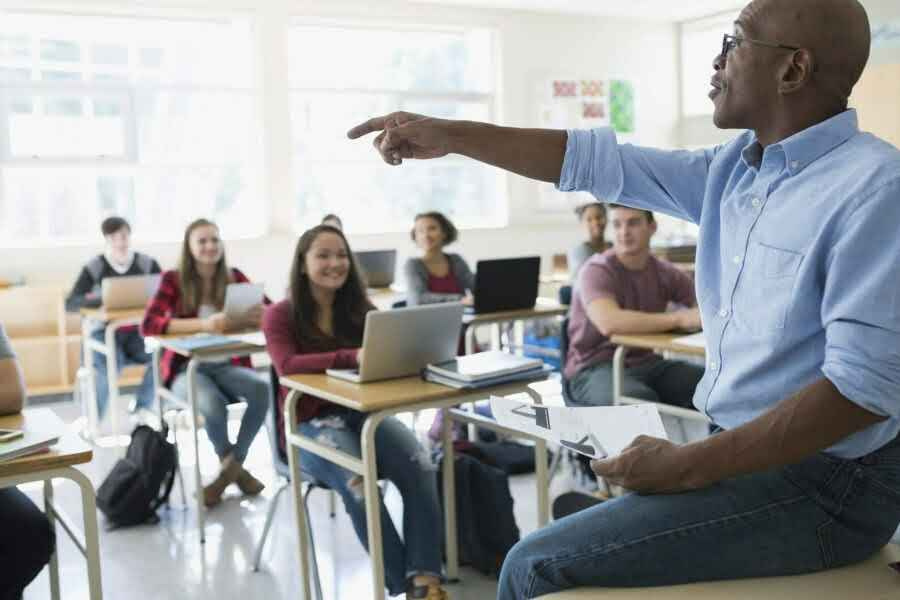 Black teacher fostering critical thinking in sixth form students, leading a lesson from the front of the classroom.