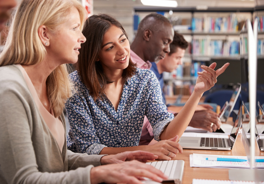 Two female teachers, one younger and one older, sit smiling at a table while they use a computer.